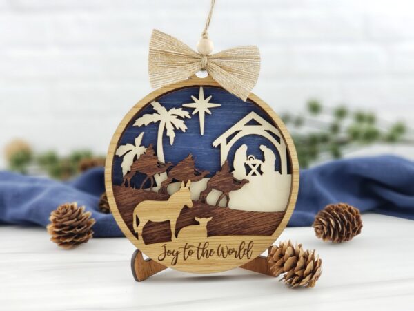 Christmas Ornaments 3D 5" Round Wood Nativity Ornament with 4 layers. Front Layer: Donkey and Lamb. Second Layer: Wise Men on Camels. Third Layer: Palm Trees, Star, Nativity Scene. Back Layer: Dark Blue. Sparkly Burlap Ribbon with wood bead and Jute twine hanger.