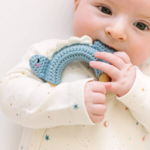 Teething baby holding a crocheted 3" wood ring with a blue stegosaurus with white round spines crocheted onto the ring.