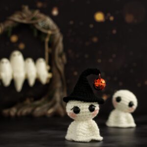 Two white crochet ghosts in a spooky setting, one wearing a witch hat with a jack o' lantern bell. Ghosts can be seen on a tree swing in the background.