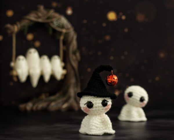 Two white crochet ghosts in a spooky setting, one wearing a witch hat with a jack o' lantern bell. Ghosts can be seen on a tree swing in the background.